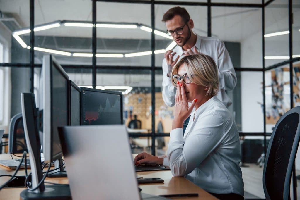 Two colleagues in an office reacting to a software issue, one seated and concerned, the other gesturing toward the screen.