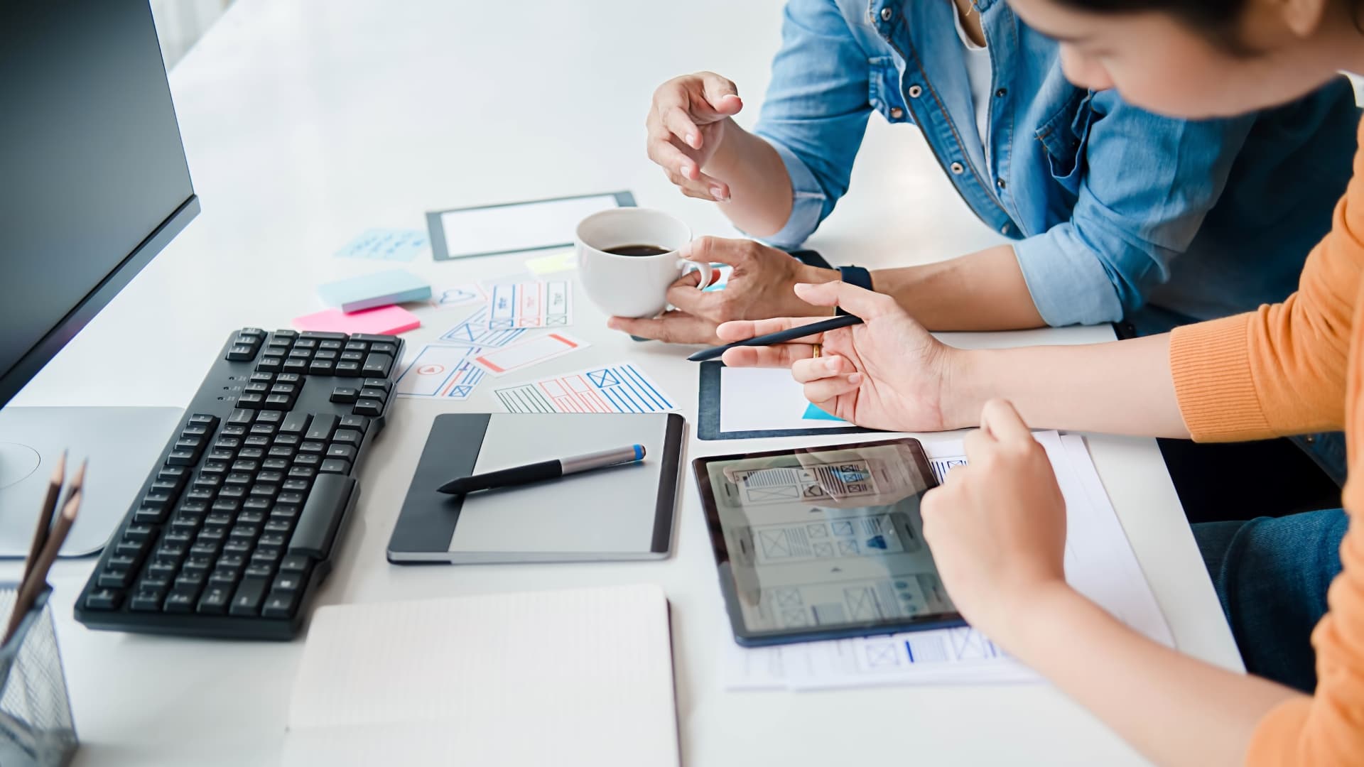 Two people collaborating at a desk with a tablet, keyboard, notebook, and wireframes, representing teamwork in performance testing planning.