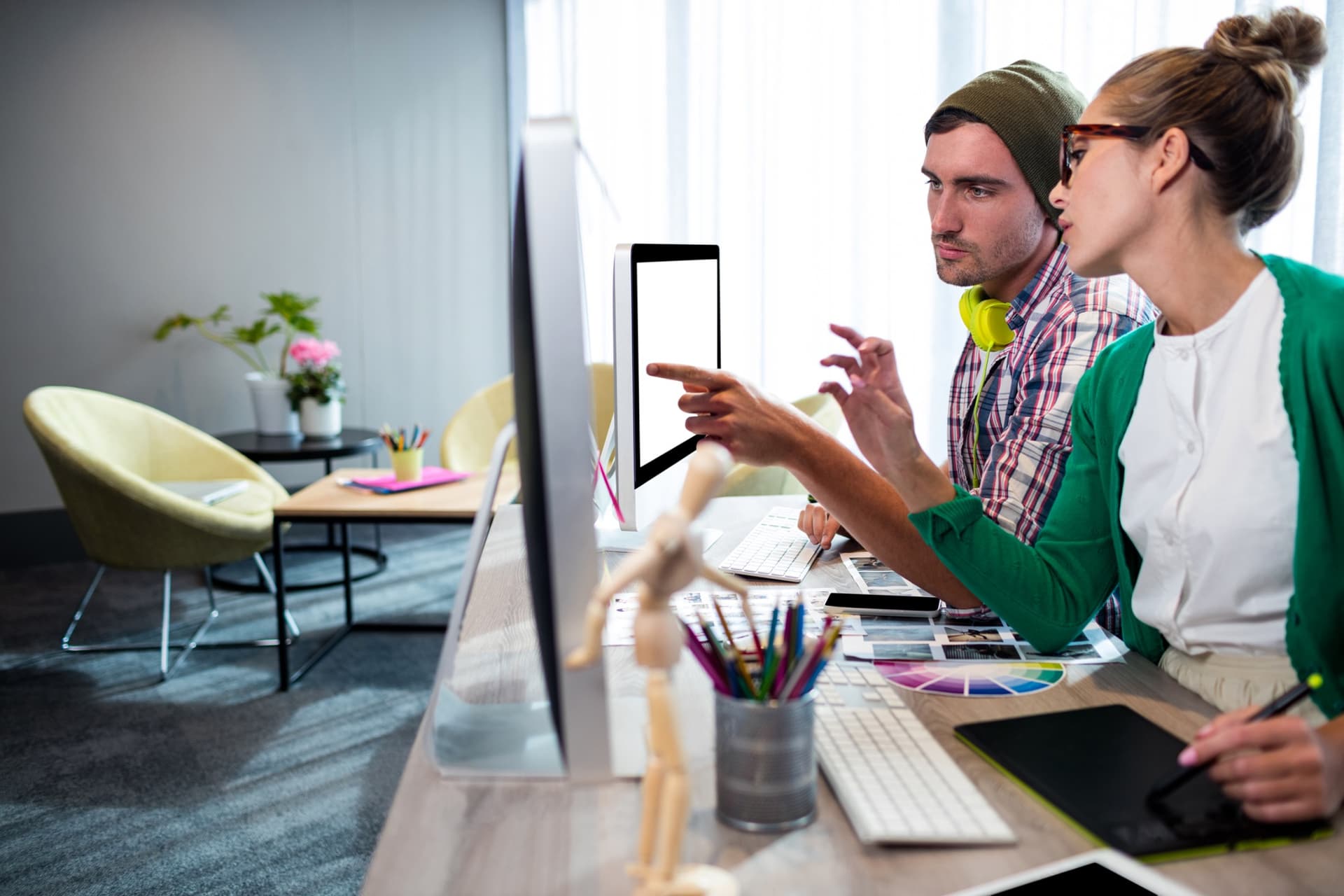 Two people collaborating at a desk with computers, color wheel, pencils, and design tools in a modern office setting.