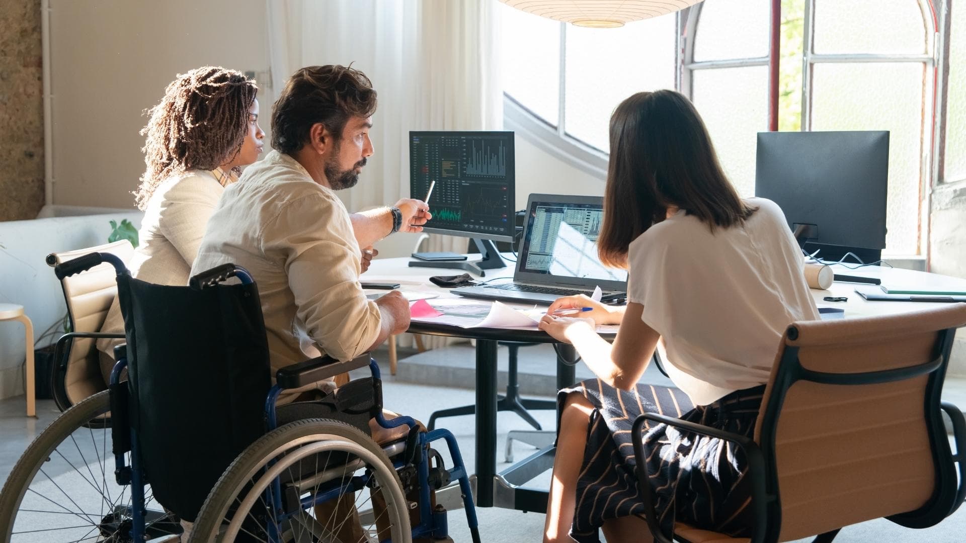 three people collaborating at a desk with multiple screens displaying data and charts, including one person using a wheelchair.