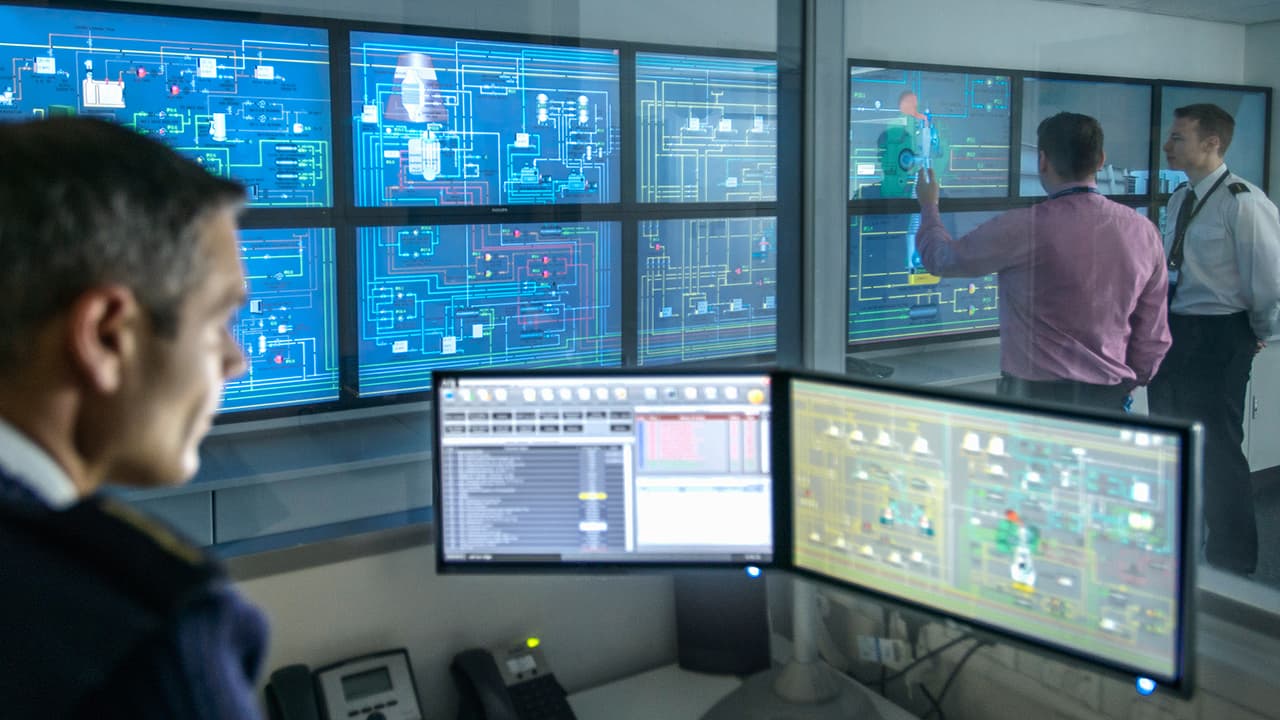 Control room with multiple monitors displaying system diagrams and data, showing a team managing and monitoring digital environments.