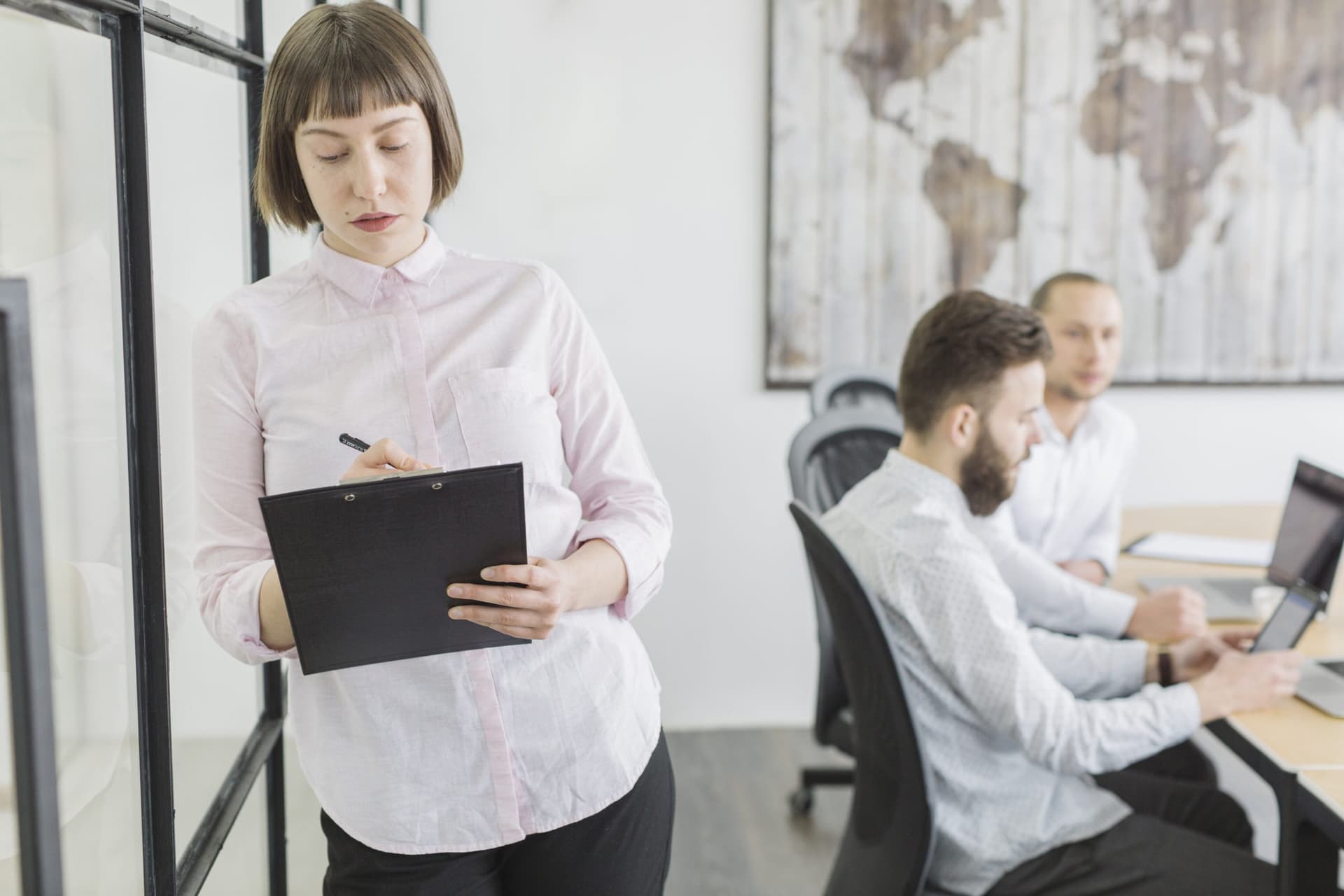 Three people in a modern office, one standing and writing on a clipboard while two others work on laptops, with a large world map on the wall in the background