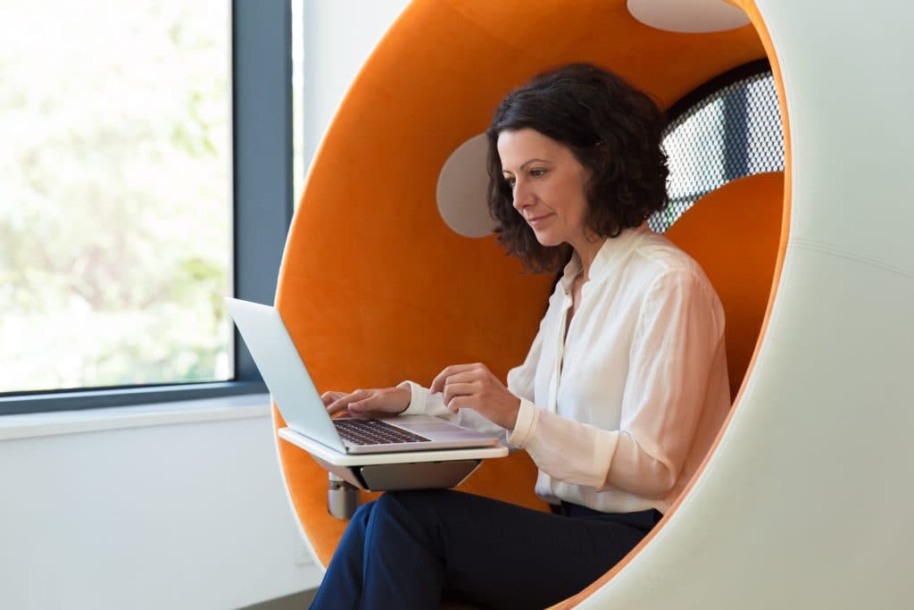 Person sitting in a modern orange pod chair, working on a laptop in a quiet and flexible learning environment.