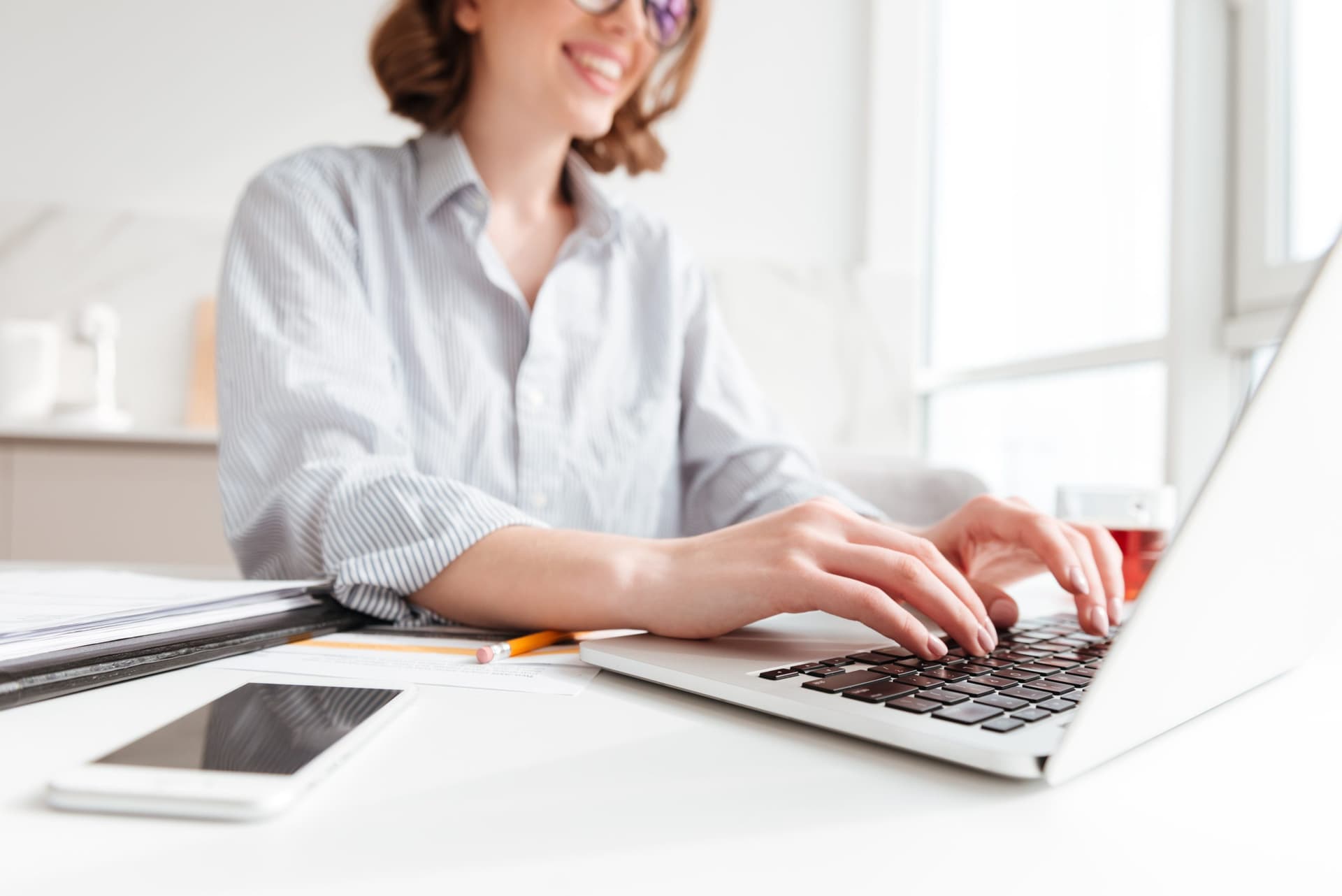 Person typing on a laptop at a desk with documents, a smartphone, and a pencil, representing digital work in a corporate environment.