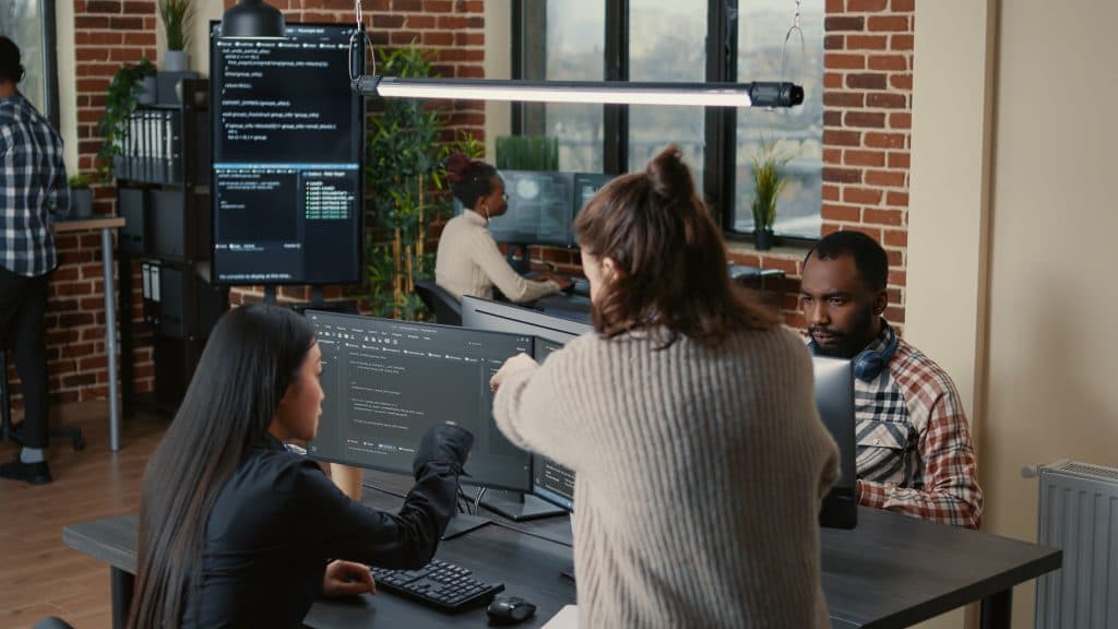 Team of professionals working at computer stations in a modern office, discussing software development and technical training.